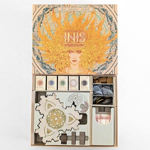 May include: Open board game box titled "Inis: Seasons of Nomad" featuring a woman with a golden headdress. The box contains game pieces, including cards, tokens, and wooden components, all neatly organized in compartments.