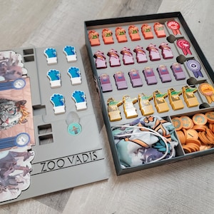 May include: A gray game board with the text "Zoo Vadis" printed on it. The board has compartments for game pieces, including blue and white animal paw prints, and a small compartment with a picture of a person. The board is open to reveal a second board with compartments for game pieces, including colorful animal figurines, ribbons, and a bag of brown tokens.