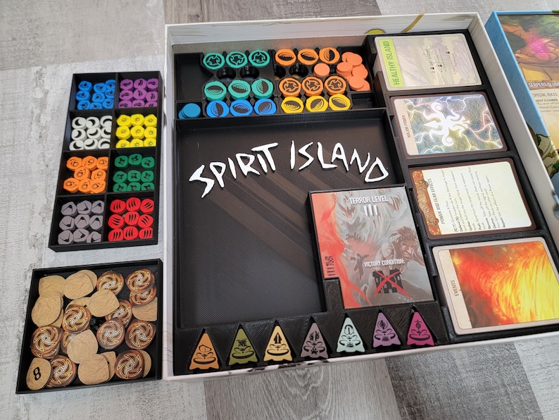 May include: A Spirit Island board game organiser with black and grey compartments for storing game pieces, cards, and tokens. The organiser has a black insert with the words "Spirit Island" printed in white.