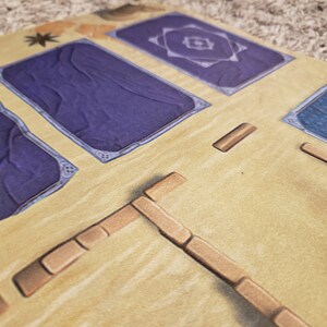 Peut inclure: Gros plan sur un plateau de jeu avec des tuiles bleues et violettes, et des pi&egrave;ces en bois brun. Le plateau est fait d'un mat&eacute;riau brun clair et a une surface textur&eacute;e.