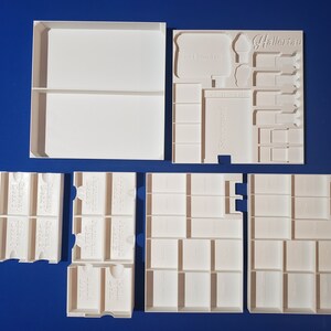 May include: White, modular storage components for board games, including trays and dividers. The components are arranged on a blue surface. Some pieces have text that reads "Hallerau" and "Game Day".