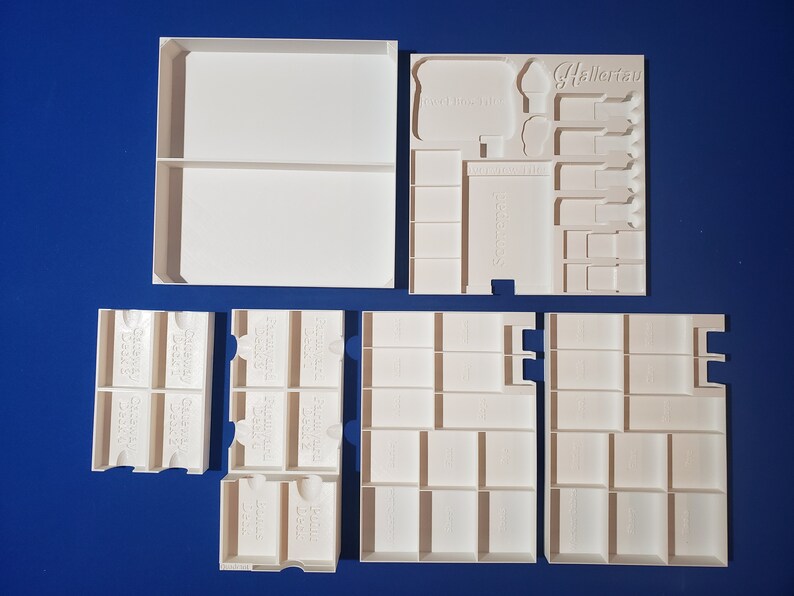 May include: White, modular storage components for board games, including trays and dividers. The components are arranged on a blue surface. Some pieces have text that reads "Hallerau" and "Game Day".