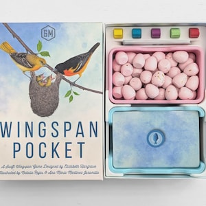 May include: Wingspan Pocket board game box and open interior. The lid features two birds at a nest and the text Wingspan Pocket. The interior contains a pink tray filled with small light pink egg tokens, a blue card deck, and five colored cubes.