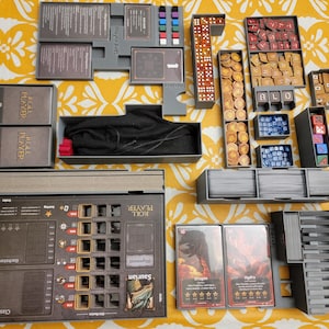 May include: A tabletop board game organizer with various components, including dice, tokens, cards, and player boards. The game pieces are arranged in gray trays and boxes. The game is set on a yellow floral patterned surface.