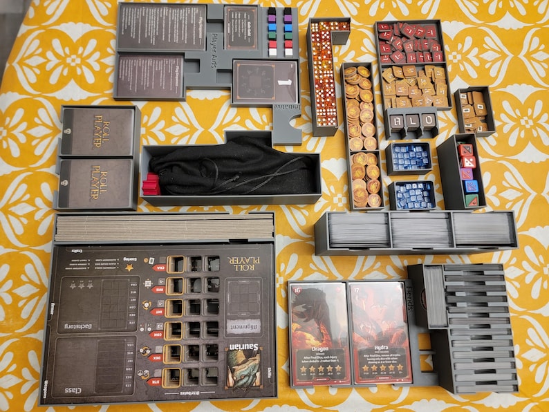 May include: A tabletop board game organizer with various components, including dice, tokens, cards, and player boards. The game pieces are arranged in gray trays and boxes. The game is set on a yellow floral patterned surface.