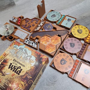 Puede incluir: Una caja de cartón marrón con el título "Defenders of the Wild" e ilustraciones de animales y personas. La caja está abierta y muestra un organizador de juegos de madera marrón con varios compartimentos y piezas de juego.