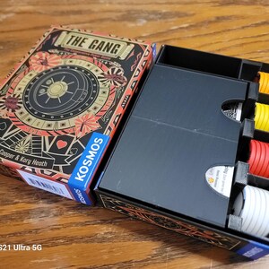 May include: A black box with a game insert for the board game "The Gang: The Cooperative Heist Game" by John Cooper & Kory Heath. The box has a black interior with dividers for organizing game components. The box is open and shows the game components, including red, yellow, and white tokens.