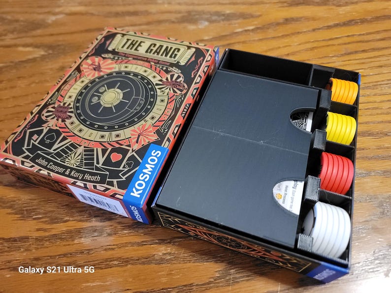 May include: A black box with a game insert for the board game "The Gang: The Cooperative Heist Game" by John Cooper & Kory Heath. The box has a black interior with dividers for organizing game components. The box is open and shows the game components, including red, yellow, and white tokens.