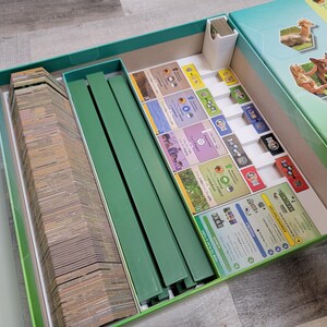 May include: Open board game box with various components. The box is teal and white, with images of animals on the lid. Inside, there are stacks of cards, green dividers, and game pieces. The game appears to be a strategy or educational game.