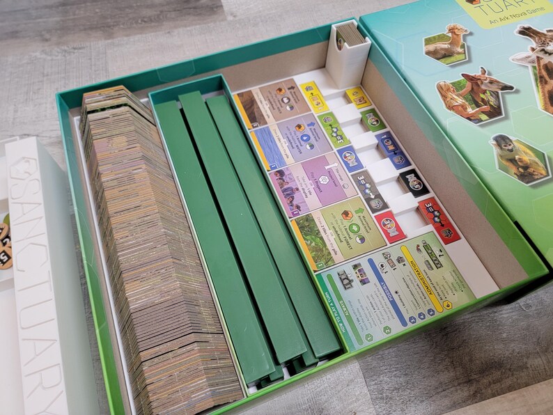 May include: Open board game box with various components. The box is teal and white, with images of animals on the lid. Inside, there are stacks of cards, green dividers, and game pieces. The game appears to be a strategy or educational game.
