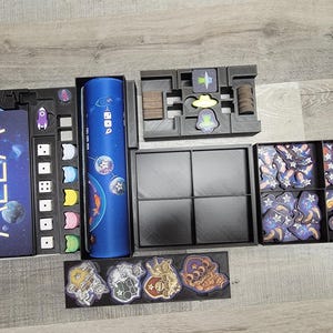 Peut inclure: Un organisateur de jeu noir et bleu avec des compartiments pour les d&eacute;s, les cartes et les jetons. L'organisateur porte l'inscription "Mlem". L'organisateur est rempli de divers composants de jeu, notamment des d&eacute;s, des cartes et des jetons.