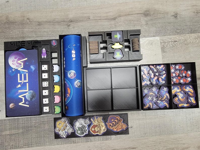 Peut inclure: Un organisateur de jeu noir et bleu avec des compartiments pour les d&eacute;s, les cartes et les jetons. L'organisateur porte l'inscription "Mlem". L'organisateur est rempli de divers composants de jeu, notamment des d&eacute;s, des cartes et des jetons.