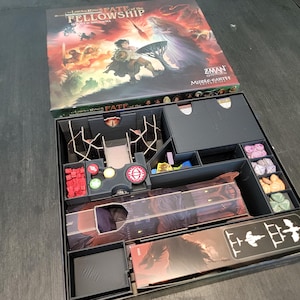 Pode incluir: Caixa de jogo de tabuleiro aberta intitulada "The Lord of the Rings: Fate of the Fellowship" com várias peças e compartimentos. A arte da caixa apresenta uma cena de fantasia com personagens e uma árvore. As peças do jogo são vermelhas, verdes, azuis e roxas.