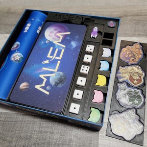 Peut inclure: Une bo&icirc;te de jeu bleue avec un design sur le th&egrave;me de l'espace. La bo&icirc;te contient des d&eacute;s, des jetons et un plateau de jeu. Le plateau de jeu pr&eacute;sente un design de galaxie avec le mot "MILLEM" imprim&eacute; dessus. La bo&icirc;te comprend &eacute;galement un ensemble de jetons color&eacute;s avec des personnages ressemblant &agrave; des chats.