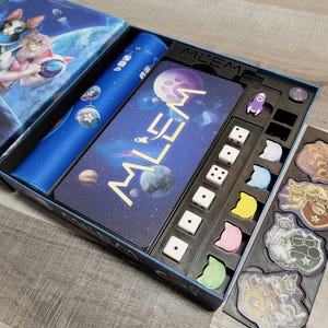 Peut inclure: Un gros plan sur un jeu de soci&eacute;t&eacute; appel&eacute; "Mlem" avec un plateau de jeu bleu et noir, des d&eacute;s et des jetons. Le plateau de jeu pr&eacute;sente un th&egrave;me spatial avec des plan&egrave;tes et des &eacute;toiles. Le jeu est con&ccedil;u pour 2 &agrave; 4 joueurs.