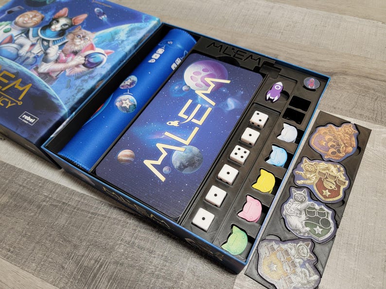 Peut inclure: Un gros plan sur un jeu de soci&eacute;t&eacute; appel&eacute; "Mlem" avec un plateau de jeu bleu et noir, des d&eacute;s et des jetons. Le plateau de jeu pr&eacute;sente un th&egrave;me spatial avec des plan&egrave;tes et des &eacute;toiles. Le jeu est con&ccedil;u pour 2 &agrave; 4 joueurs.
