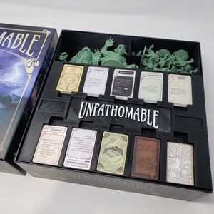 Könnte beinhalten: Eine schwarze Box mit einem Schaumstoffeinsatz zum Organisieren von Spielkomponenten. Die Box trägt den Schriftzug "Unfathomable" auf der Vorderseite. Der Schaumstoffeinsatz verfügt über Fächer für Karten, Miniaturen und andere Spielteile.