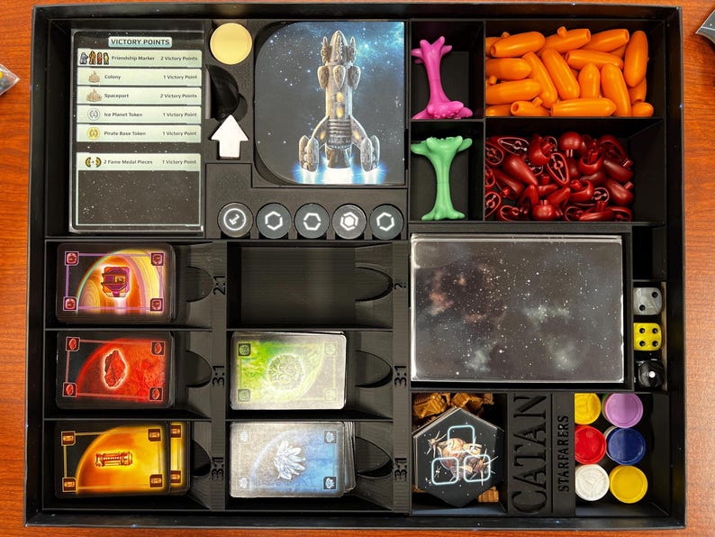 May include: A Catan: Starfarers game organizer insert with compartments for cards, tokens, and other game components. The insert is made of black plastic and has a clear plastic lid. The insert is labeled with the game's name and logo.