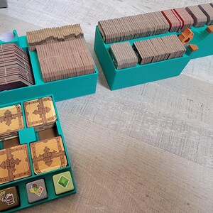 May include: Teal-colored game organizer with multiple compartments. Contains stacks of cards, wooden tokens, and miniature treasure chests. The organizer is designed to hold game components, keeping them organized and accessible.