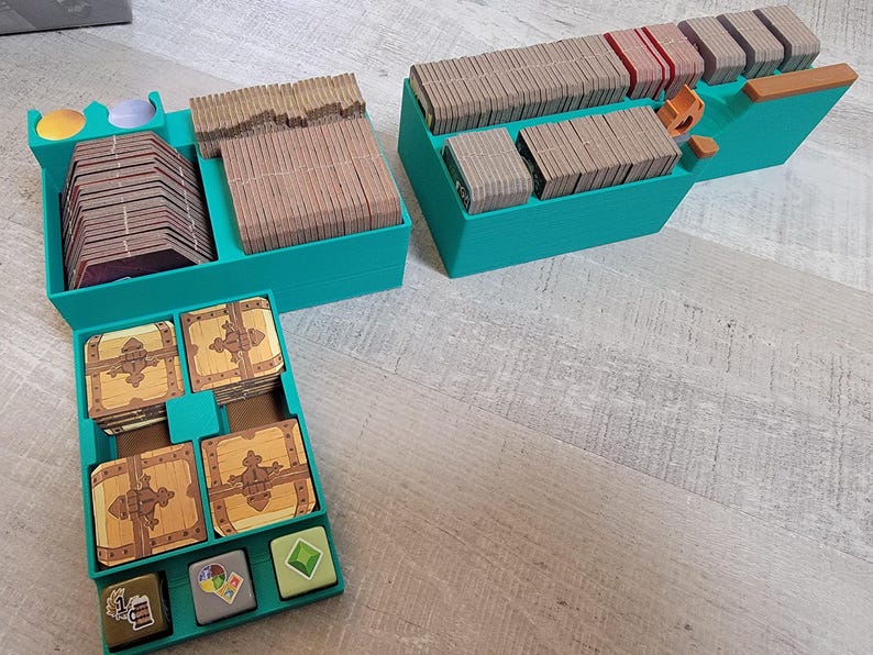 May include: Teal-colored game organizer with multiple compartments. Contains stacks of cards, wooden tokens, and miniature treasure chests. The organizer is designed to hold game components, keeping them organized and accessible.