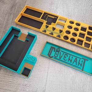 May include: Three-piece game organizer set in teal, yellow, and brown. The set includes a rectangular tray with the word "COVENANT", a teal and brown tray with the word "Solo", and a yellow tray with various compartments for game pieces.
