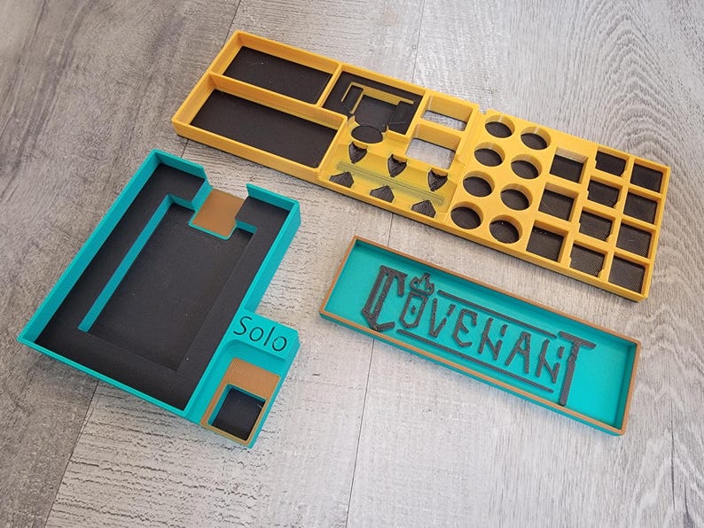 May include: Three-piece game organizer set in teal, yellow, and brown. The set includes a rectangular tray with the word "COVENANT", a teal and brown tray with the word "Solo", and a yellow tray with various compartments for game pieces.