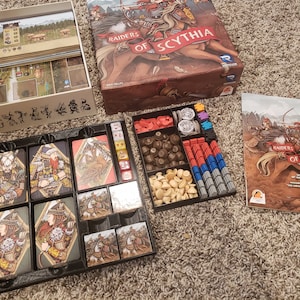 Könnte beinhalten: Ein schwarzer Plastik-Organizer mit Fächern zum Verstauen von Spielsteinen und Karten. Der Organizer ist mit verschiedenen Spielkomponenten gefüllt, darunter Würfel, Spielsteine und Karten. Die Spielbox befindet sich im Hintergrund, mit dem Titel "Raiders of Scythia" auf der Vorderseite.