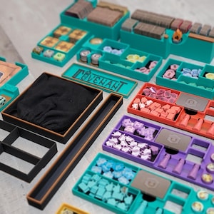 May include: A collection of teal, black, brown, red, purple, and yellow game organizers. The organizers are filled with game pieces, including dice, tokens, and cards. The word "COVENANT" is visible on a teal container.