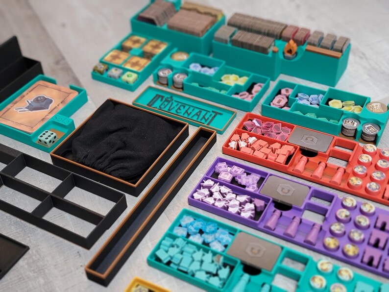 May include: A collection of teal, black, brown, red, purple, and yellow game organizers. The organizers are filled with game pieces, including dice, tokens, and cards. The word "COVENANT" is visible on a teal container.