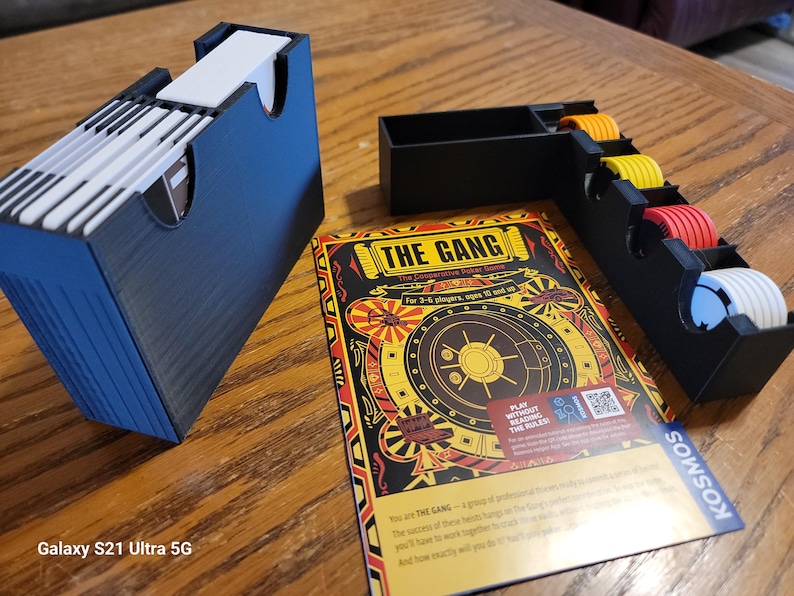 May include: A black and yellow board game called "The Gang" with a safe on the cover. The game is for 3-8 players, ages 10 and up. The game includes a black card holder with white cards inside, a black tray with yellow, orange, and red tokens, and a black tray with white tokens.