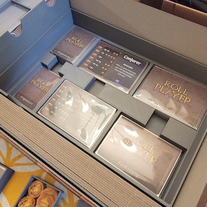 May include: An open board game box with various components. The box is divided into compartments holding cards, game pieces, and wooden sticks. The cards are labeled "Roll Player" and "Conjurer". The box is gray and brown.