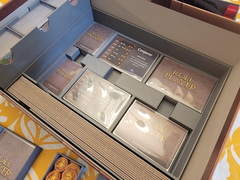 May include: An open board game box with various components. The box is divided into compartments holding cards, game pieces, and wooden sticks. The cards are labeled "Roll Player" and "Conjurer". The box is gray and brown.