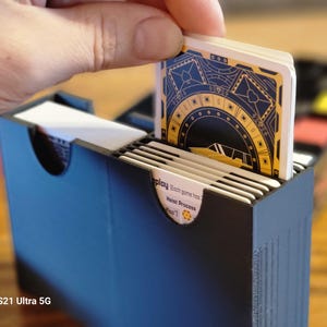 May include: A black card game box with a deck of cards being inserted. The cards have a gold and black design with a circular pattern and the text "Replay. Each game comes from Host Process [Rear]" is visible on the bottom card.