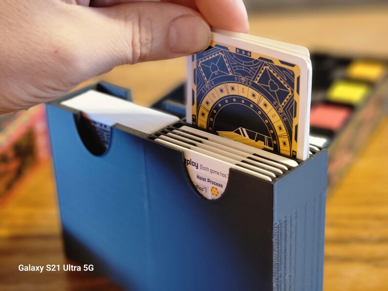 May include: A black card game box with a deck of cards being inserted. The cards have a gold and black design with a circular pattern and the text "Replay. Each game comes from Host Process [Rear]" is visible on the bottom card.