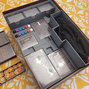 May include: A board game organizer with compartments for cards, tokens, and player aids. The box is gray and brown, with various game components visible, including cards, dice, and a black bag. The box is open, revealing the organized contents.
