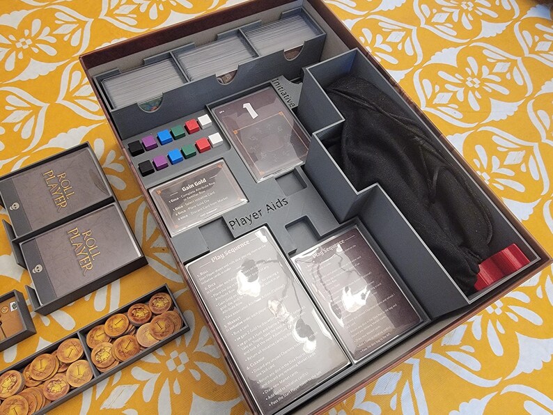 May include: A board game organizer with compartments for cards, tokens, and player aids. The box is gray and brown, with various game components visible, including cards, dice, and a black bag. The box is open, revealing the organized contents.