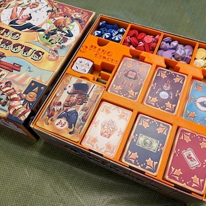 Op de afbeelding: Een 3 Ring Circus bordspel insert met oranje scheidingswanden en compartimenten. De insert bevat een verscheidenheid aan spelstukken, waaronder dobbelstenen, tokens en kaarten. De kaarten bevatten kleurrijke illustraties van circusartiesten.