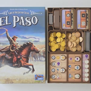 May include: Open board game box titled "Great Western Trail El Paso." The box art depicts a cowboy on horseback roping cattle. Inside, brown plastic trays hold game pieces, including cards, tokens, and yellow coin-like pieces.