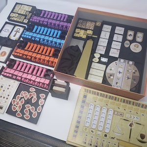 May include: A game organizer for the board game "The Great Zimbabwe". The organizer includes multiple compartments for storing the game's components, including wooden pieces, cards, and tiles. The organizer is made of black plastic and has a brown cardboard box.