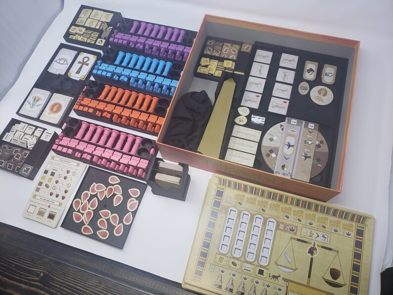 May include: A game organizer for the board game "The Great Zimbabwe". The organizer includes multiple compartments for storing the game's components, including wooden pieces, cards, and tiles. The organizer is made of black plastic and has a brown cardboard box.