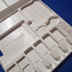 May include: A white, 3D-printed game organizer with compartments for game components. The organizer has text that reads "Jewel Box Tiles", "Overview Tiles", "Scorepad", and "Hallertau". The design features various shapes and sizes for different game pieces.