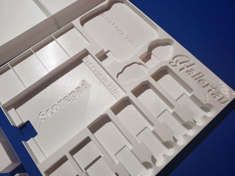 May include: A white, 3D-printed game organizer with compartments for game components. The organizer has text that reads "Jewel Box Tiles", "Overview Tiles", "Scorepad", and "Hallertau". The design features various shapes and sizes for different game pieces.