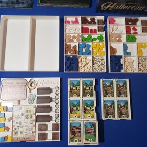 May include: A board game with various components, including wooden pieces in multiple colors, cards, and white storage trays. The game's title, "Hallertau," is visible on a game board element. The components are arranged on a blue surface.