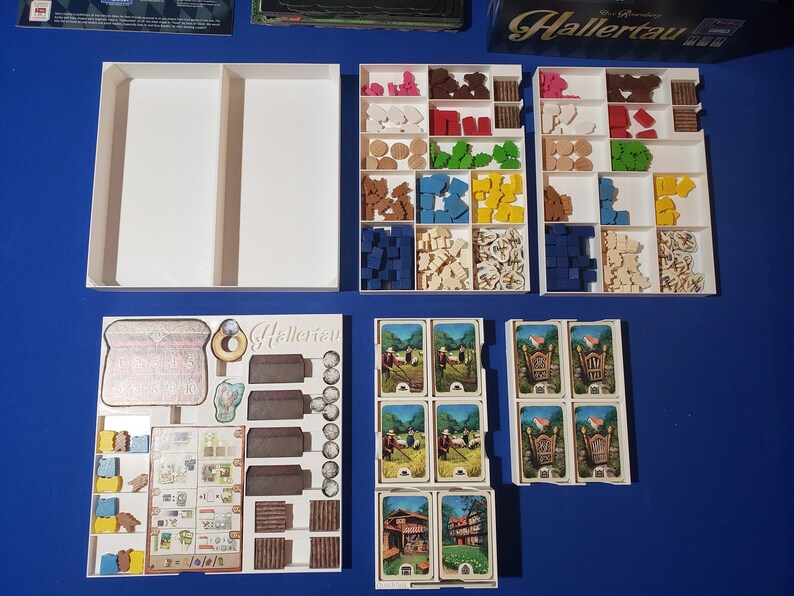 May include: A board game with various components, including wooden pieces in multiple colors, cards, and white storage trays. The game's title, "Hallertau," is visible on a game board element. The components are arranged on a blue surface.