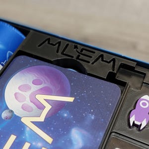 Peut inclure: Gros plan sur une bo&icirc;te de jeu avec une fus&eacute;e violette et une carte avec une plan&egrave;te et des &eacute;toiles. La bo&icirc;te porte l'inscription "Mlem".