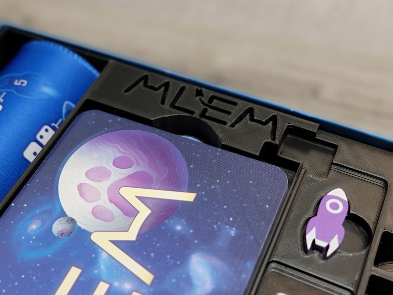 Peut inclure: Gros plan sur une bo&icirc;te de jeu avec une fus&eacute;e violette et une carte avec une plan&egrave;te et des &eacute;toiles. La bo&icirc;te porte l'inscription "Mlem".