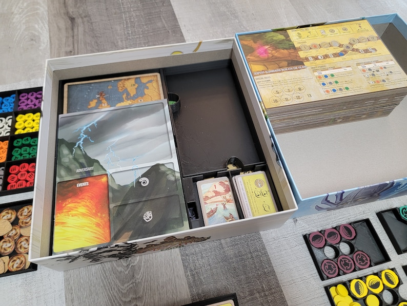 May include: A white box with a black insert for organising board game components. The box contains a map, cards, and tokens. The box is open and the contents are visible. The box is sitting on a wooden table.