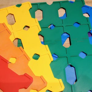 May include: A fan of colorful, interlocking plastic tiles in a rainbow of hues: red, orange, yellow, green, blue, and purple. Each tile has a unique shape with hexagonal and rectangular cutouts, suggesting a building toy or puzzle.