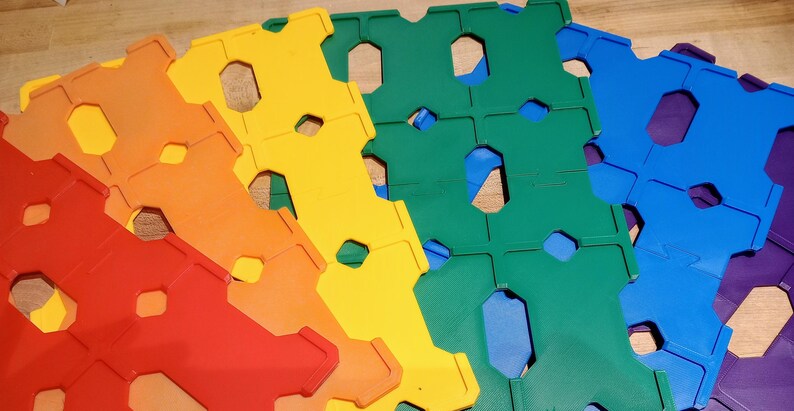 May include: A fan of colorful, interlocking plastic tiles in a rainbow of hues: red, orange, yellow, green, blue, and purple. Each tile has a unique shape with hexagonal and rectangular cutouts, suggesting a building toy or puzzle.
