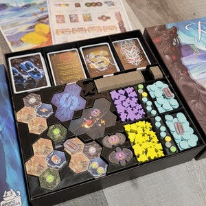 May include: A black organiser box with compartments for storing game pieces and cards. The box is filled with various coloured plastic pieces, including blue, purple, yellow, and green. The box is labelled "Rorvic Call the Abyss".